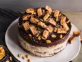 Eine köstliche Snickers Torte mit Schokoladenglasur, Erdnüssen und vielen Snickers-Stücken obendrauf.