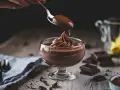Ein Löffel mit cremigem Mousse au Chocolat tropft auf eine kunstvoll aufgespritze Portion in einem Glas.