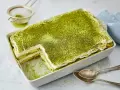 Ein cremiges Matcha Tiramisu, bestäubt mit feinem grünen Pulver, wartet in einer Auflaufform darauf, genossen zu werden. Löffel liegen bereit.