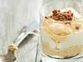 Cremiger cheesecake im glas, geschichtet mit weißer Creme und garniert mit zerbröselten Keksen. Ein Löffel liegt daneben.