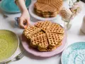 Ein Stapel herzförmiger Waffeln ohne Ei auf einem rosa Teller, bereit zum Genießen. Daneben liegen bunte Teller und eine Gabel.