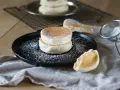 Zwei fluffige japanische Pancakes auf einem schwarzen Teller, bestäubt mit Puderzucker. Daneben ein Stück Pancake und ein Sieb.