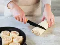 Hände schneiden Teigrolle für Zimtschnecken. Bereits geformte Schnecken warten in einer Pfanne auf das Backen. Ein tolles Zimtschnecken Rezept!