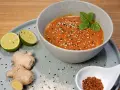 Schüssel mit cremiger Erdnusssauce, garniert mit Sesam und Minze. Daneben Limetten, Ingwer und Chiliflocken – Zutaten für ein leckeres Erdnusssauce Rezept.