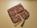 Ein rechteckiger Zwetschgenkuchen mit Streuseln, ein Stück wird mit einem Kuchenheber herausgehoben.