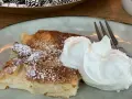 Ein Stück saftiger Apfelkuchen, bestäubt mit Puderzucker, serviert mit einer großen Portion Schlagsahne und einer Gabel.
