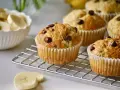 Frisch gebackene schoko bananen muffins mit Schokostückchen auf einem Gitter. Daneben eine Schüssel mit Bananenscheiben.