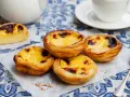 Vier köstliche Pastel de Nata auf einem blauen Tischtuch, mit einer Tasse Kaffee im Hintergrund.