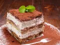 Ein Stück cremiges Tiramisu ohne Ei, bestäubt mit Kakao und garniert mit Minze, auf einem Teller mit Kakaopulver und einer Gabel.