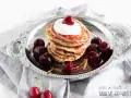 Ein Stapel Bananen Pancakes mit Kirschen und Sahne auf einem Silbertablett, garniert mit frischen Kirschen.
