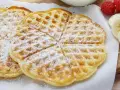 Zuckerbestäubte Buttermilchwaffeln, die auf einem Holzbrett liegen. Daneben sind Himbeeren und Bananenscheiben zu sehen.