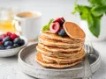 Ein Stapel veganer Pancakes, garniert mit Kirschen und Blaubeeren, daneben eine Schale Beeren und eine Tasse Kaffee.