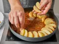 Hände legen Apfelspalten in eine Pfanne mit karamellisierter Butter. Die Zubereitung der klassischen Tarte Tatin beginnt.