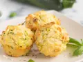 Drei goldbraune Zucchini-Muffins mit Kräutern und Käse auf einem Teller, garniert mit Basilikum.