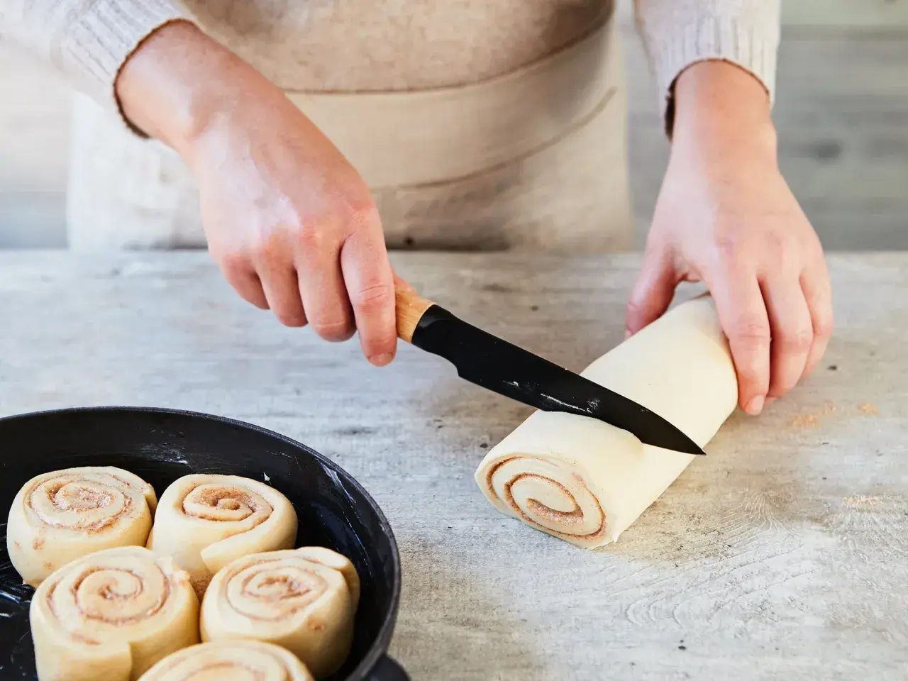 Hände schneiden Teigrolle für Zimtschnecken. Bereits geformte Schnecken warten in einer Pfanne auf das Backen. Ein tolles Zimtschnecken Rezept!