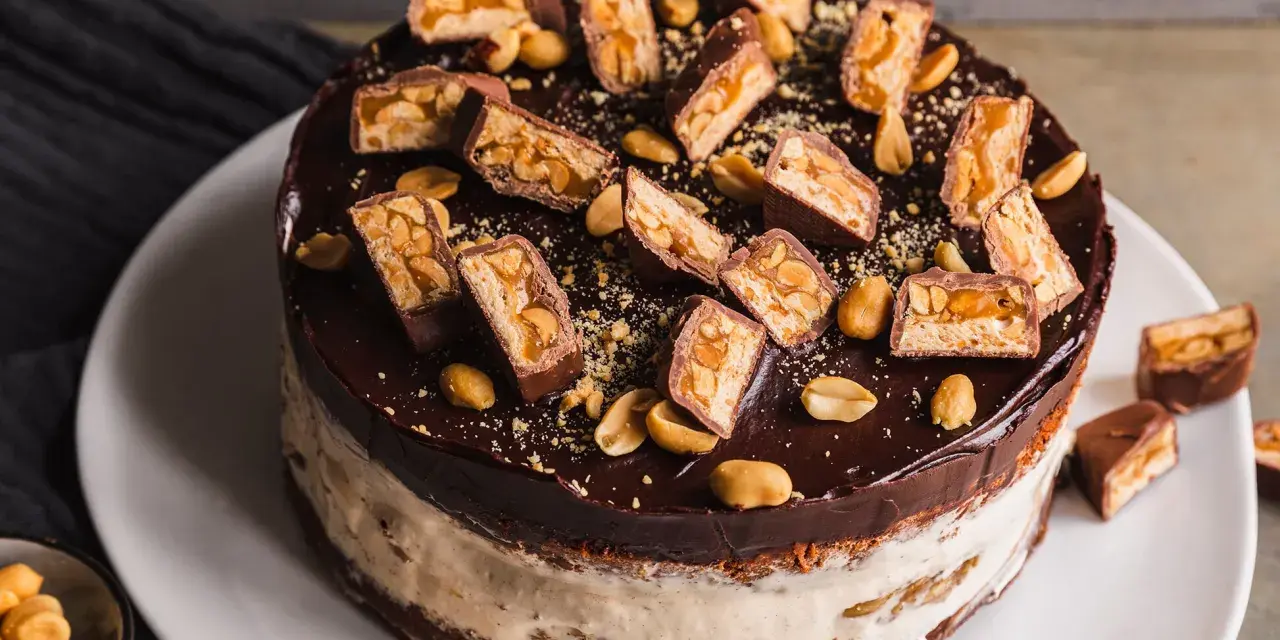 Eine köstliche Snickers Torte mit Schokoladenglasur, Erdnüssen und vielen Snickers-Stücken obendrauf.