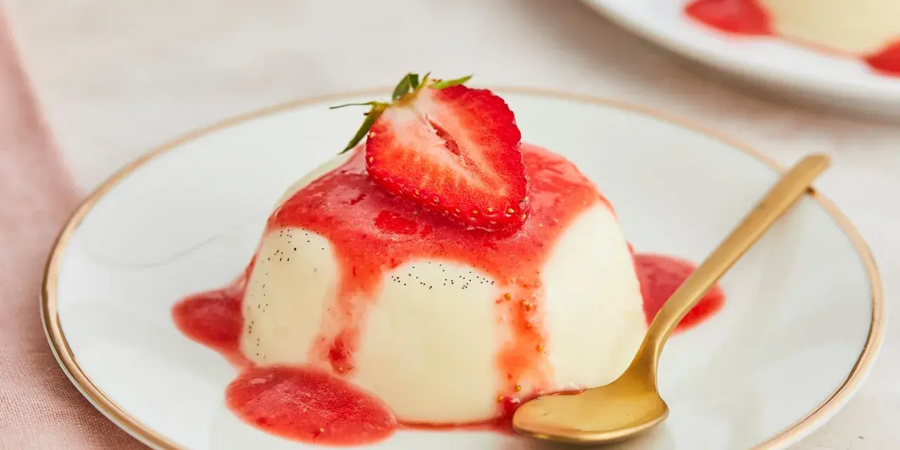 Cremige Panna Cotta mit Erdbeersauce und frischer Erdbeere, garniert mit Vanillepunkten. Eine goldene Löffel liegt daneben.