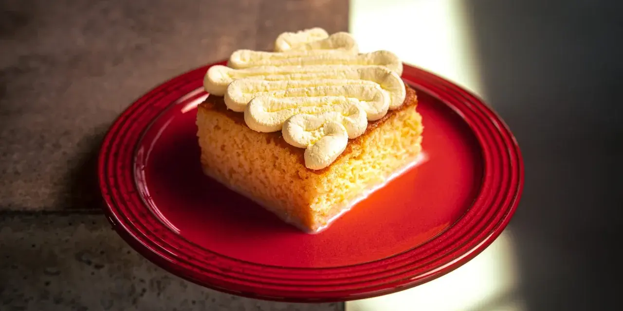 Ein Stück saftiger Tres Leches Kuchen mit Sahnetupfern auf einem roten Teller, bereit zum Genießen.