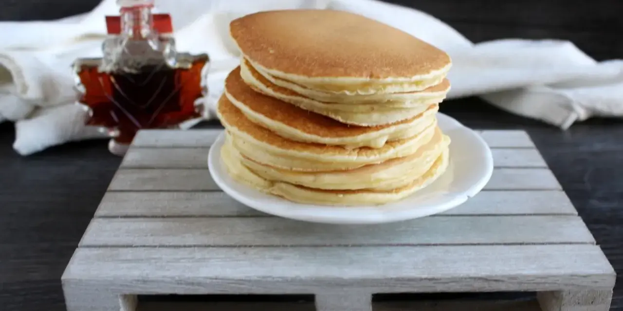 Ein Stapel goldbrauner Pfannkuchen, bereit für ein köstliches Frühstück. Daneben eine kleine Flasche Ahornsirup. Ein einfaches Pfannkuchen Rezept für jeden Tag.