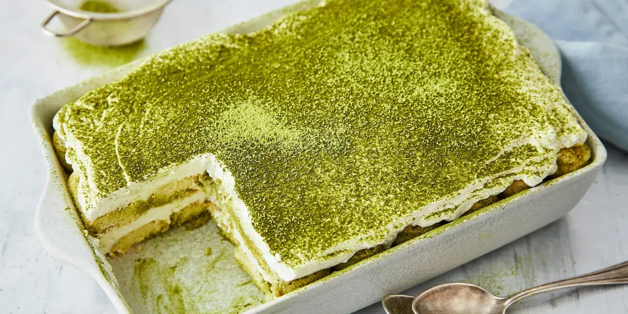 Ein cremiges Matcha Tiramisu, bestäubt mit feinem grünen Pulver, wartet in einer Auflaufform darauf, genossen zu werden. Löffel liegen bereit.