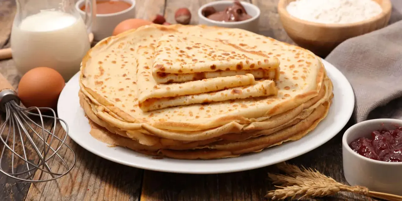 Ein Stapel goldbrauner Pfannkuchen, zubereitet nach einem sehr guten Pfannkuchen Rezept, daneben Zutaten wie Milch, Eier, Mehl, Marmelade und Nuss-Nougat-Creme.