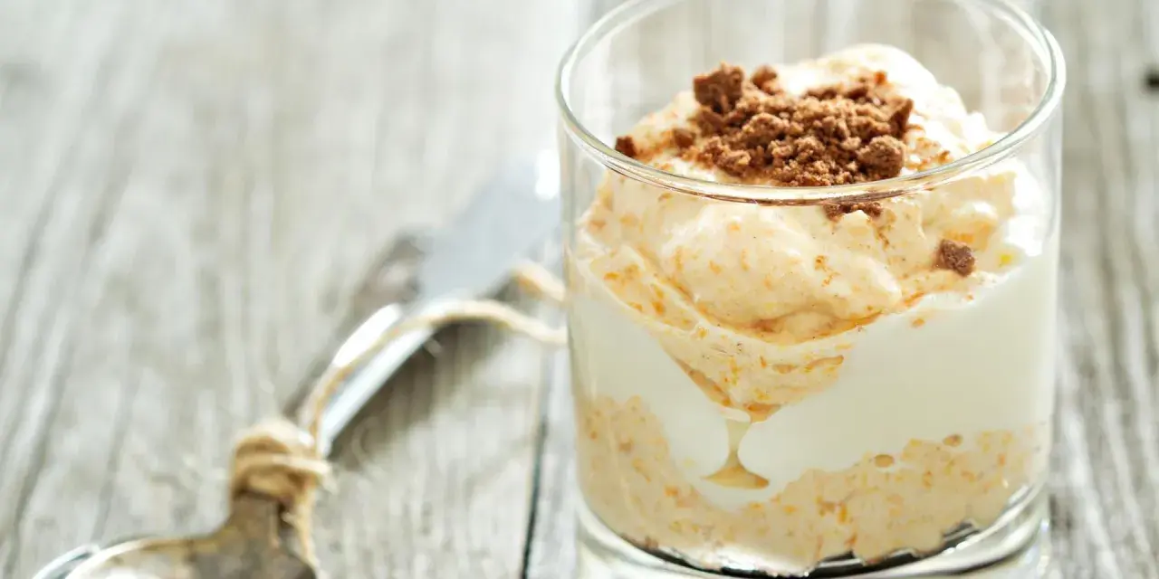 Cremiger cheesecake im glas, geschichtet mit weißer Creme und garniert mit zerbröselten Keksen. Ein Löffel liegt daneben.