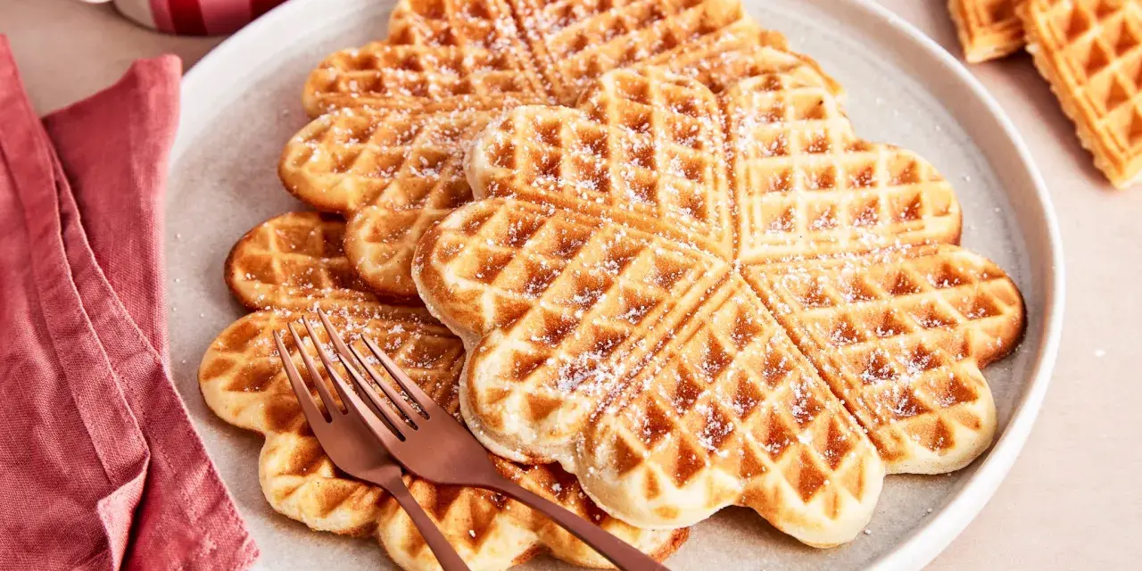 Herzförmige Waffeln, bestäubt mit Puderzucker. Ein köstlicher waffelteig ohne butter, serviert auf einem Teller mit zwei Gabeln.