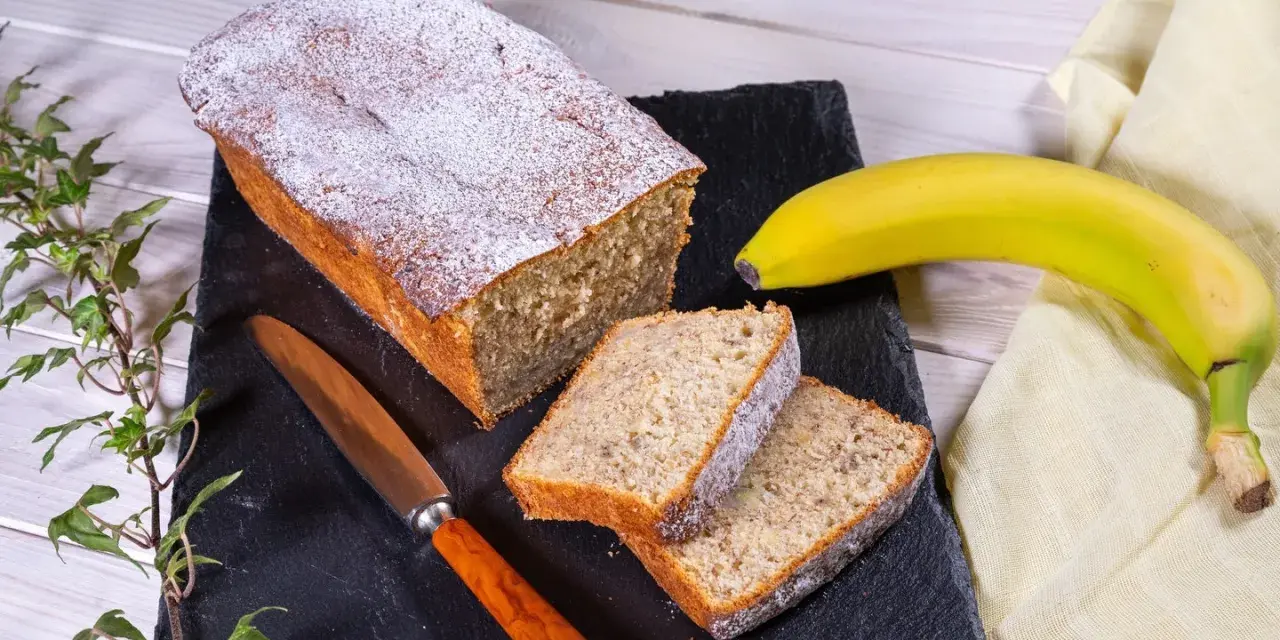 Saftiger Bananenkuchen, bestäubt mit Puderzucker, neben einer Banane und einem Messer. Perfekt für ein Bananenkuchen Rezept.