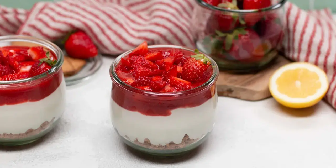 Zwei Gläser mit cremigem Erdbeer-Cheesecake, garniert mit frischen Erdbeeren und Erdbeersauce. Daneben ein Glas voller Erdbeeren und eine Zitronenhälfte.