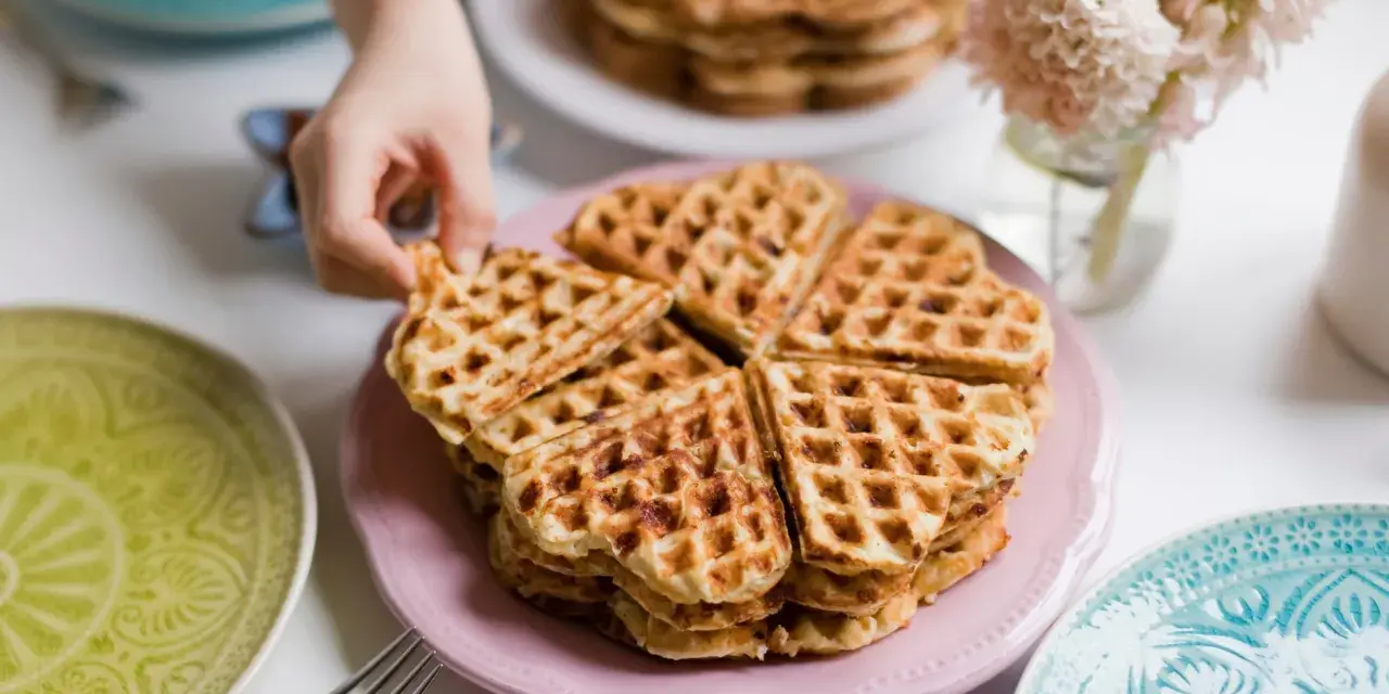 Ein Stapel herzförmiger Waffeln ohne Ei auf einem rosa Teller, bereit zum Genießen. Daneben liegen bunte Teller und eine Gabel.