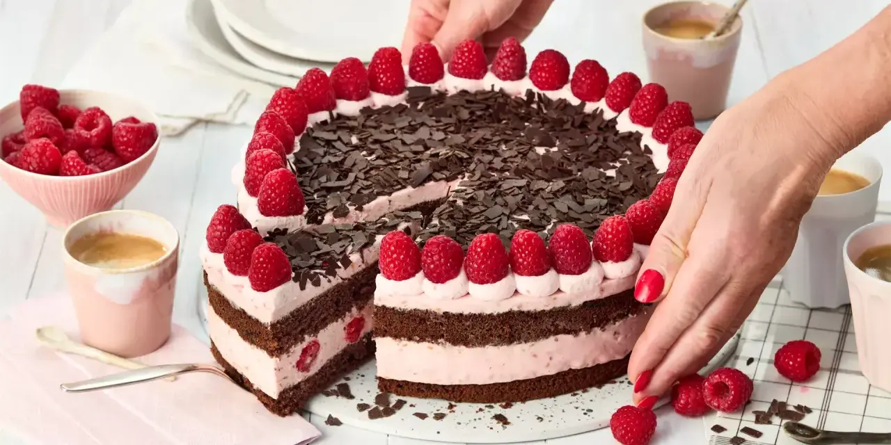 Eine köstliche Schoko Himbeer Torte, verziert mit frischen Himbeeren und Schokostreuseln, wird von Händen arrangiert.