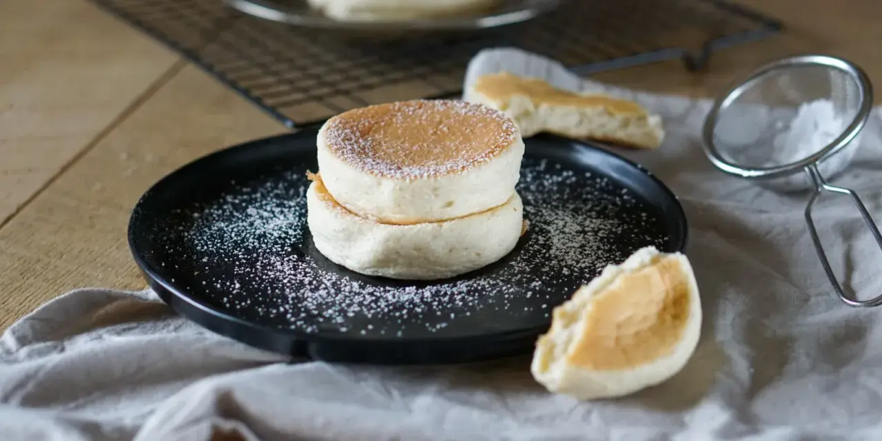 Zwei fluffige japanische Pancakes auf einem schwarzen Teller, bestäubt mit Puderzucker. Daneben ein Stück Pancake und ein Sieb.