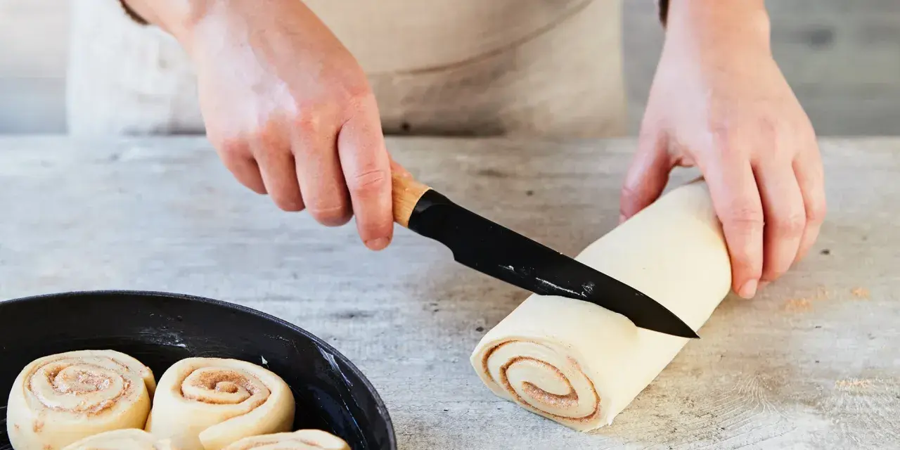 Hände schneiden Teigrolle für Zimtschnecken. Bereits geformte Schnecken warten in einer Pfanne auf das Backen. Ein tolles Zimtschnecken Rezept!