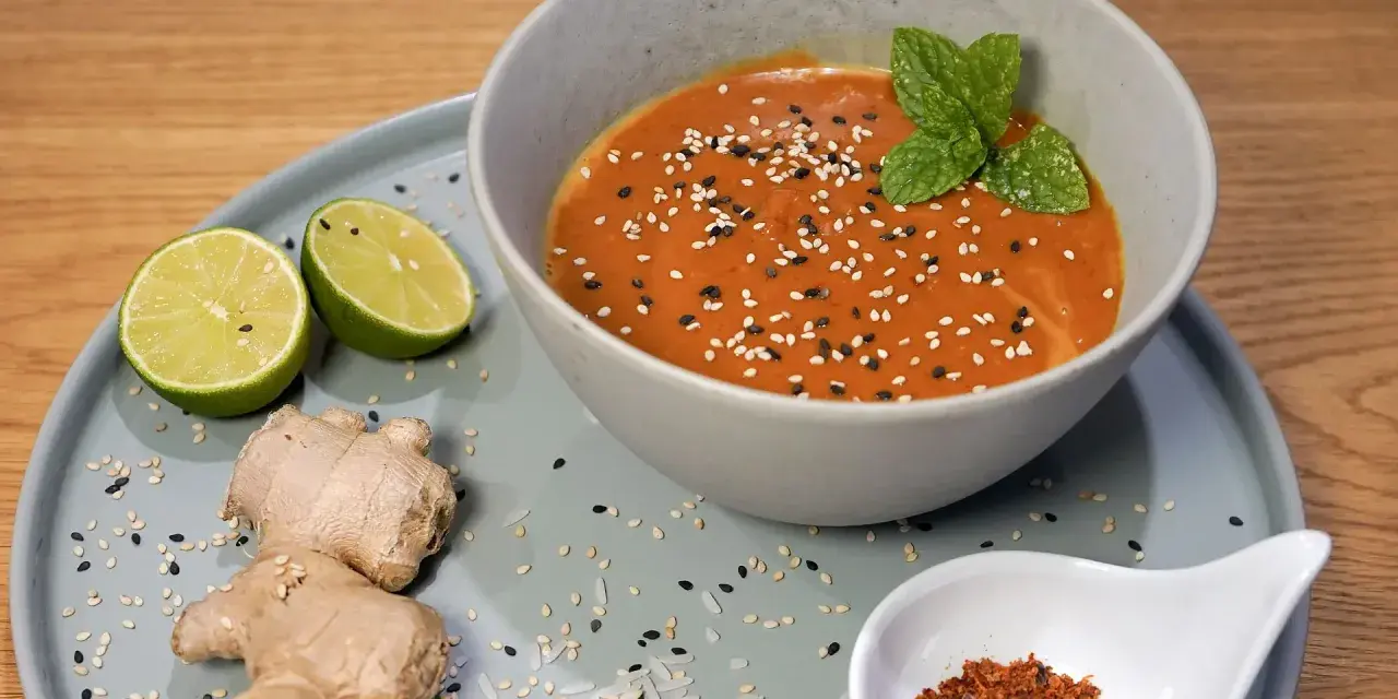 Schüssel mit cremiger Erdnusssauce, garniert mit Sesam und Minze. Daneben Limetten, Ingwer und Chiliflocken – Zutaten für ein leckeres Erdnusssauce Rezept.