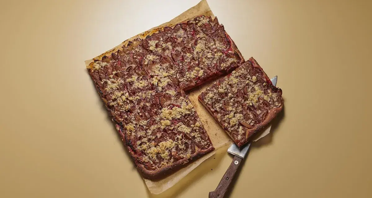 Ein rechteckiger Zwetschgenkuchen mit Streuseln, ein Stück wird mit einem Kuchenheber herausgehoben.