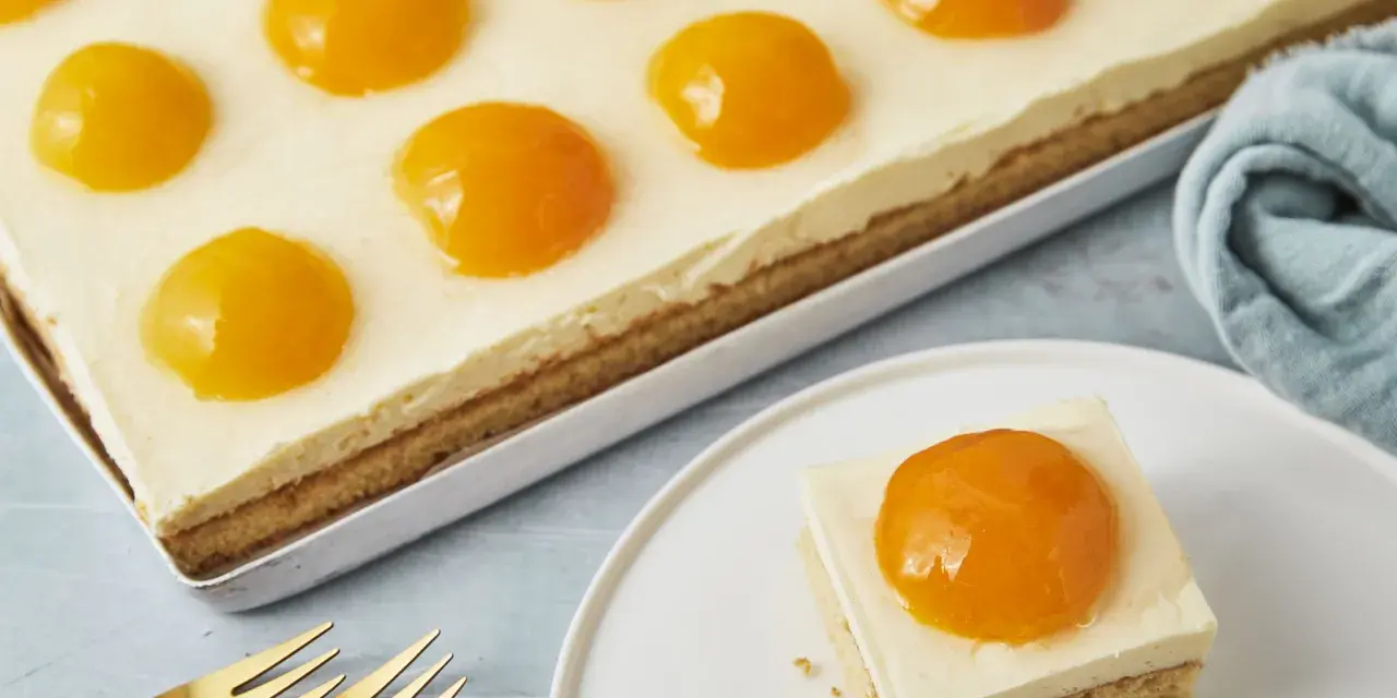 Ein rechteckiger Kuchen mit einer cremigen Schicht und vielen Pfirsichhälften, die wie ein Spiegelei Kuchen aussehen. Ein Stück auf einem Teller daneben.
