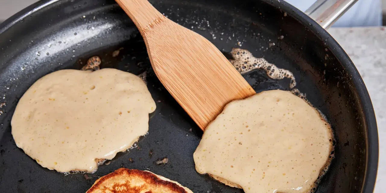 Buttermilch pancakes braten in der Pfanne. Ein Holzspatel wendet einen goldbraunen Pancake.