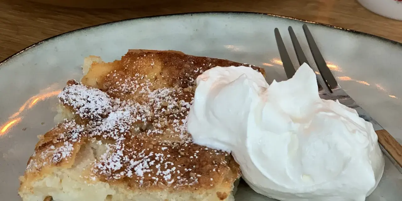 Ein Stück saftiger Apfelkuchen, bestäubt mit Puderzucker, serviert mit einer großen Portion Schlagsahne und einer Gabel.