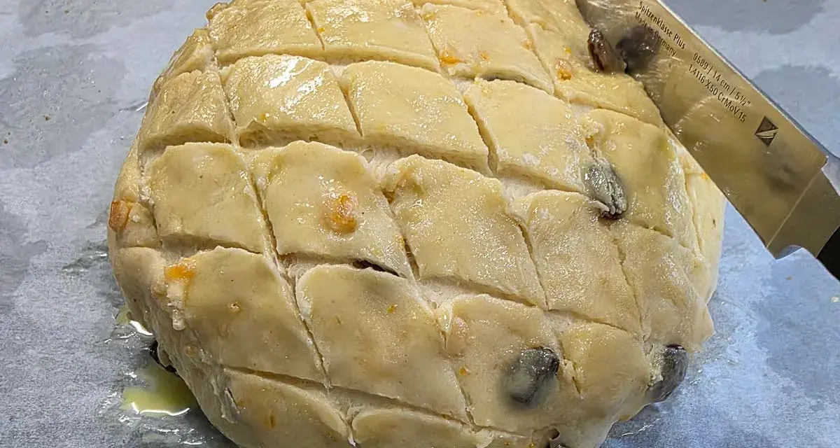 Ein runder Teigling mit eingeschnittenen Rauten, bereit für das Osterfladen Rezept. Rosinen und Orangenschale sind sichtbar.