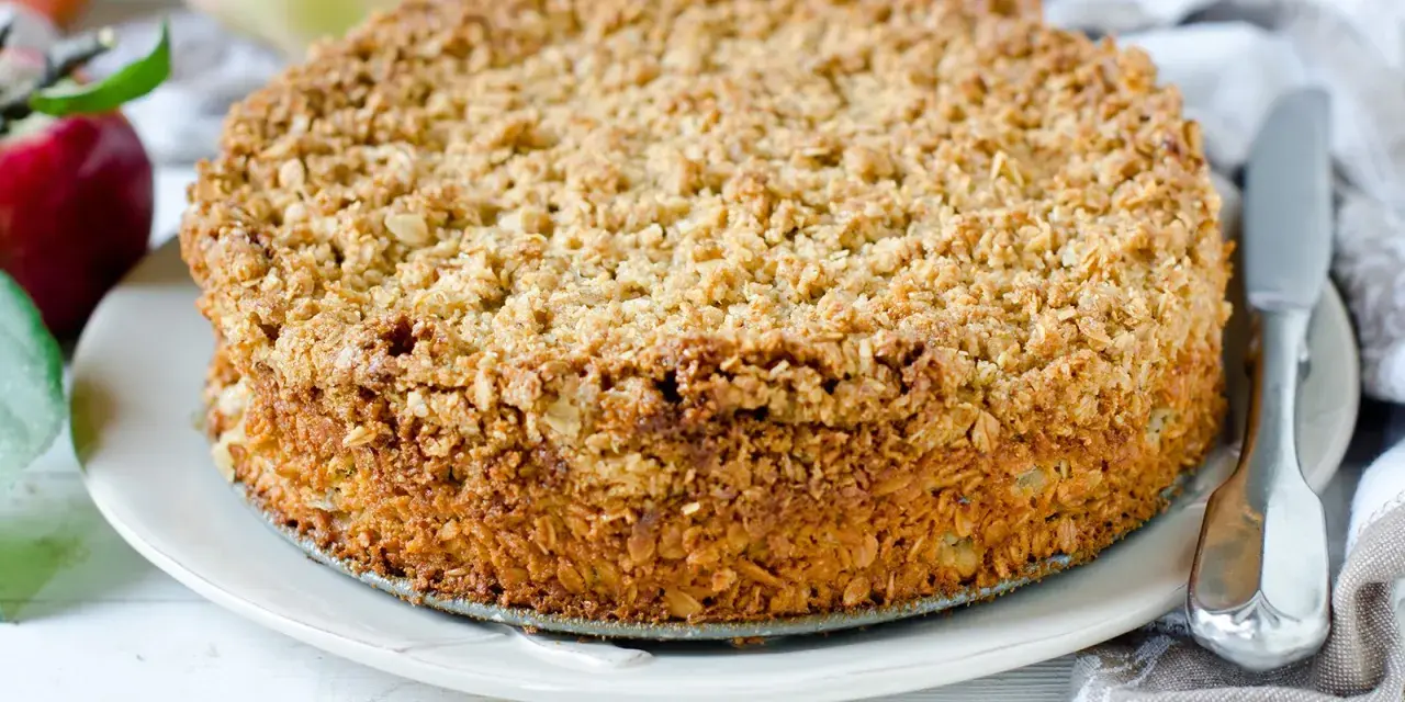 Ein goldbrauner Haferflocken Kuchen mit knuspriger Kruste, daneben Äpfel und ein Messer.