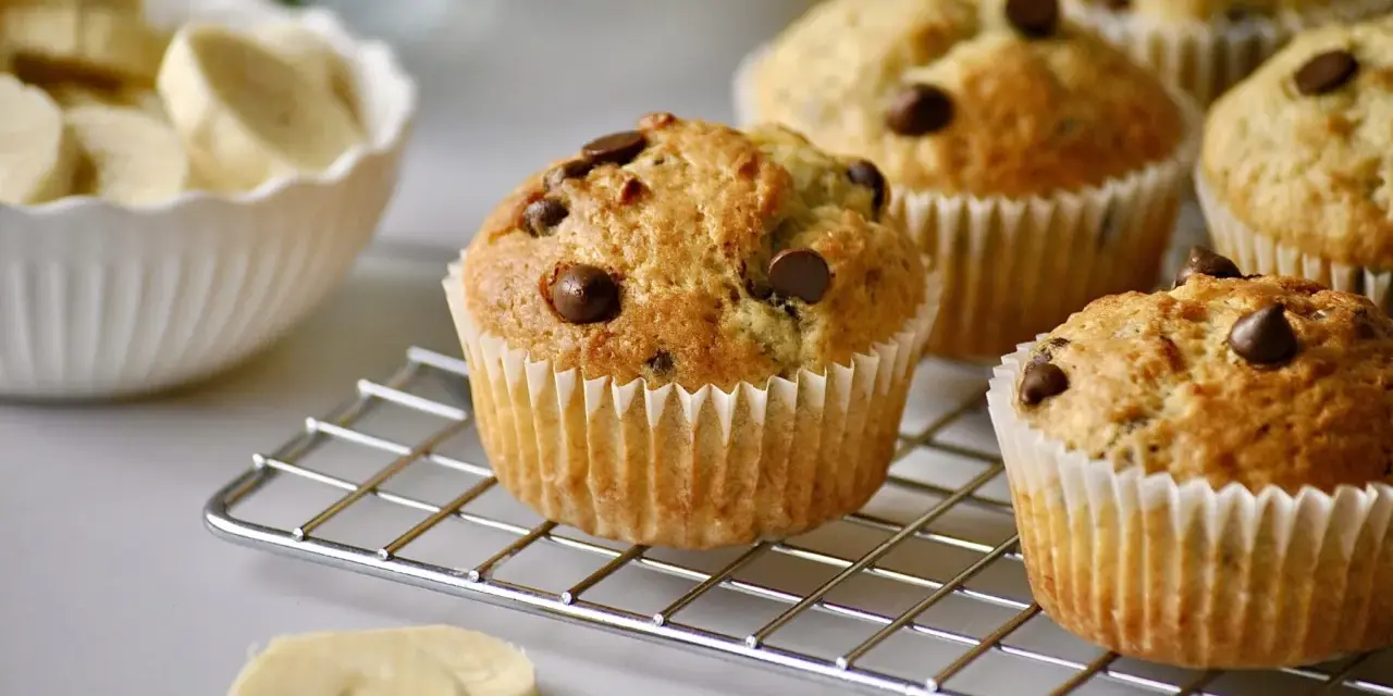 Frisch gebackene schoko bananen muffins mit Schokostückchen auf einem Gitter. Daneben eine Schüssel mit Bananenscheiben.