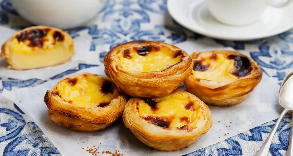 Vier köstliche Pastel de Nata auf einem blauen Tischtuch, mit einer Tasse Kaffee im Hintergrund.