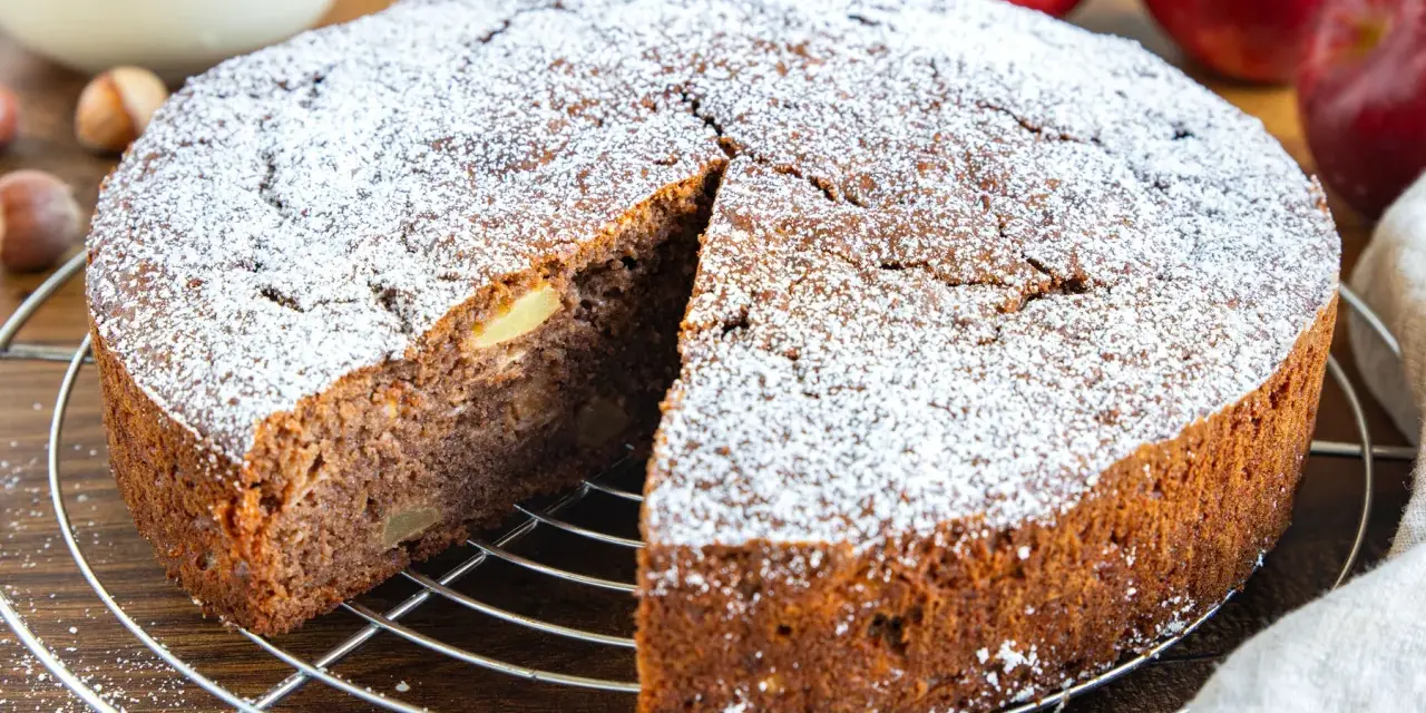 Ein saftiger russischer Apfelkuchen, bestäubt mit Puderzucker, wartet auf seine Genießer. Apfelstücke sind im dunklen Teig sichtbar.