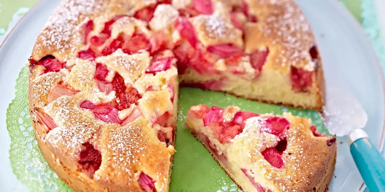 Ein saftiger Erdbeer Rhabarber Kuchen, bestäubt mit Puderzucker, liegt auf einem Teller. Ein Stück wurde bereits entnommen.