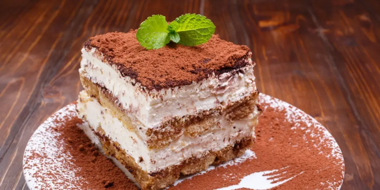 Ein Stück cremiges Tiramisu ohne Ei, bestäubt mit Kakao und garniert mit Minze, auf einem Teller mit Kakaopulver und einer Gabel.