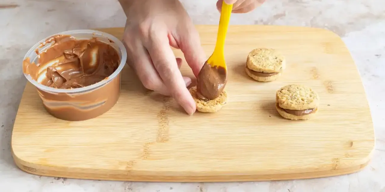 Hände belegen Kekse mit Nutella. Leckere Nutella Kekse entstehen auf dem Holzbrett.