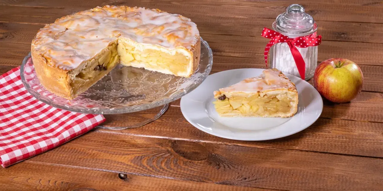 Omas weltbester Apfelkuchen, saftig und mit Zuckerguss überzogen, wartet auf einem Tortenständer. Ein Stück auf einem Teller und ein Apfel daneben laden zum Genießen ein.
