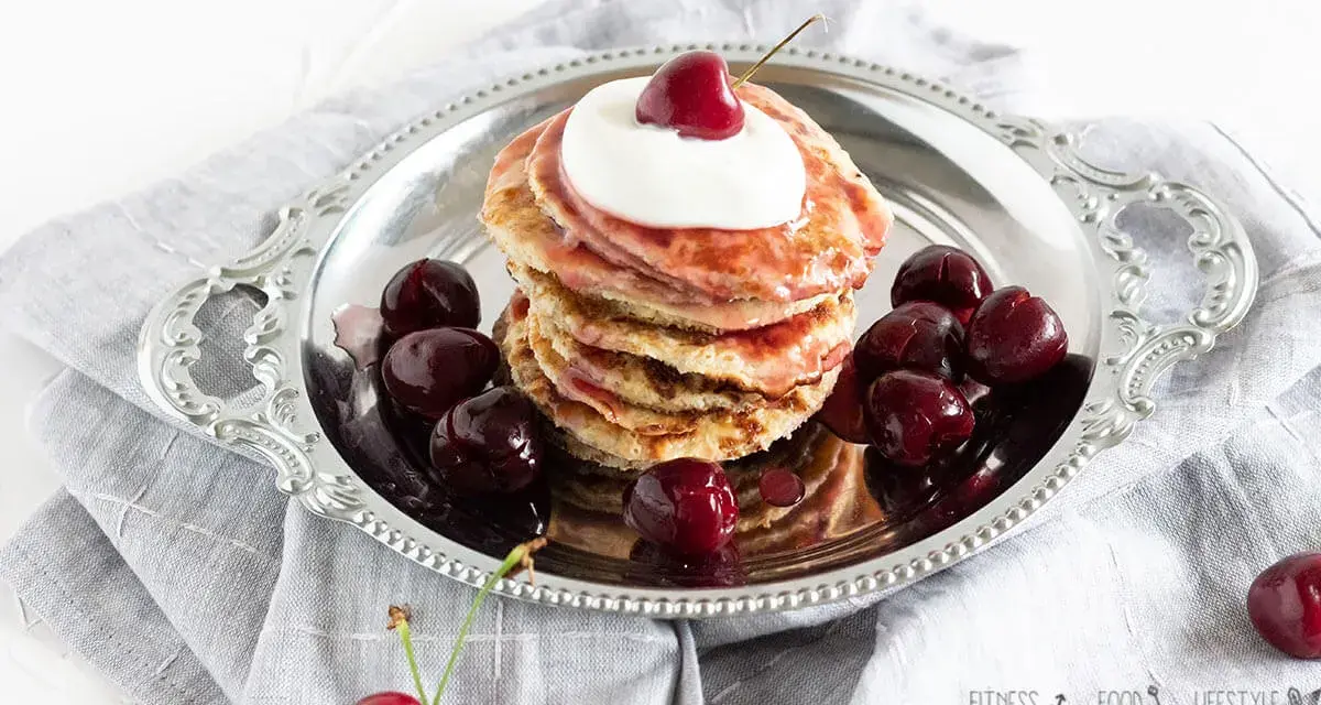Ein Stapel Bananen Pancakes mit Kirschen und Sahne auf einem Silbertablett, garniert mit frischen Kirschen.
