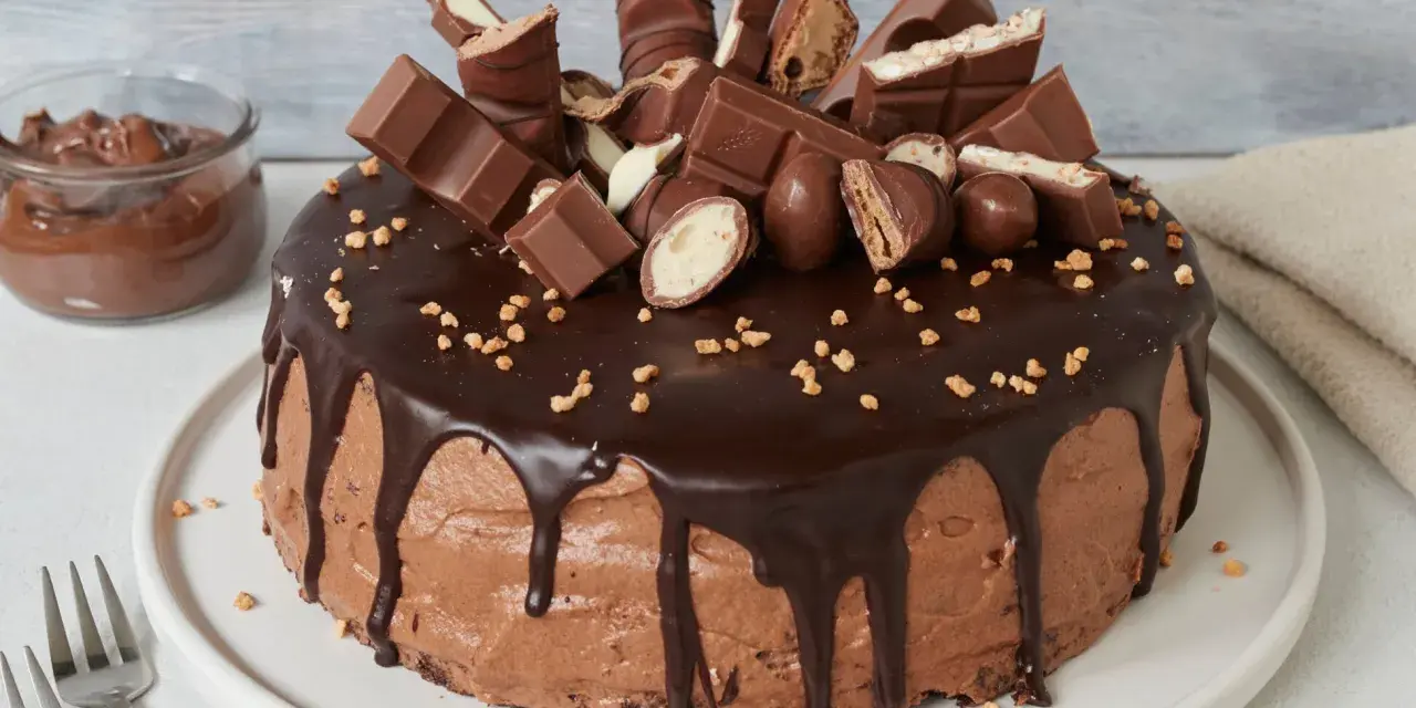 Eine üppige Nutella Torte, dekoriert mit vielen Schokoriegeln und Pralinen, daneben ein Glas Nutella.