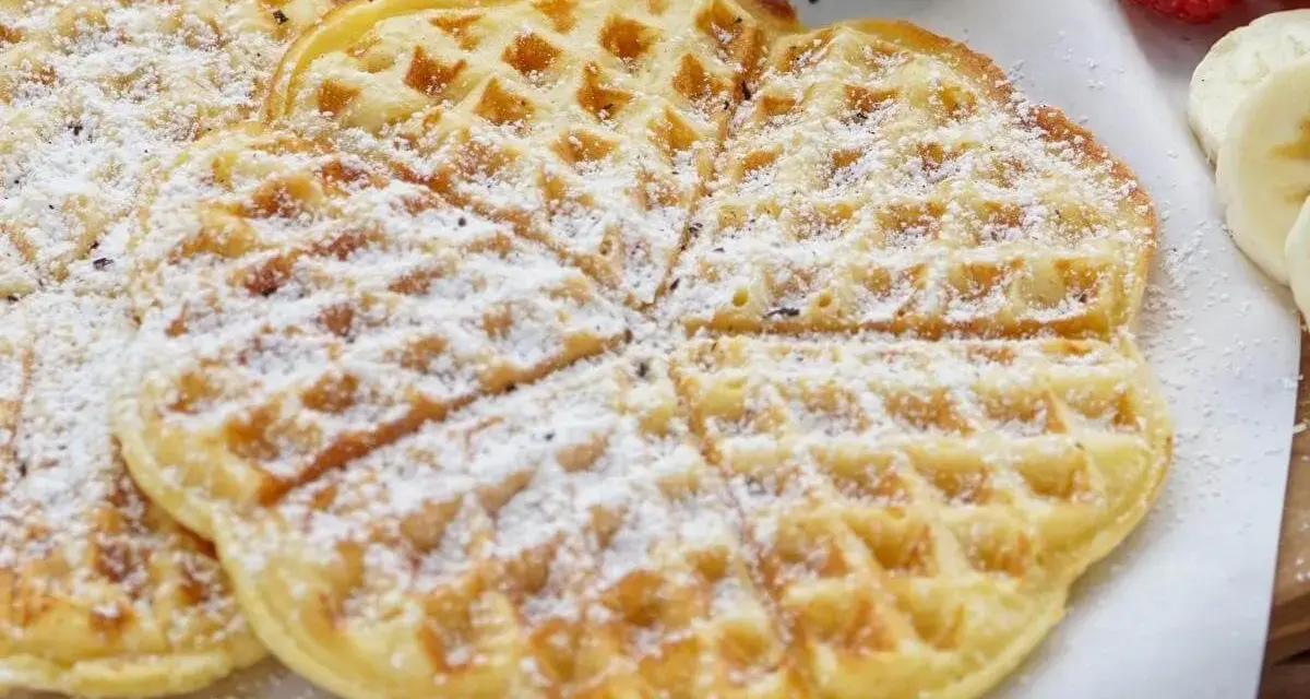 Zuckerbestäubte Buttermilchwaffeln, die auf einem Holzbrett liegen. Daneben sind Himbeeren und Bananenscheiben zu sehen.